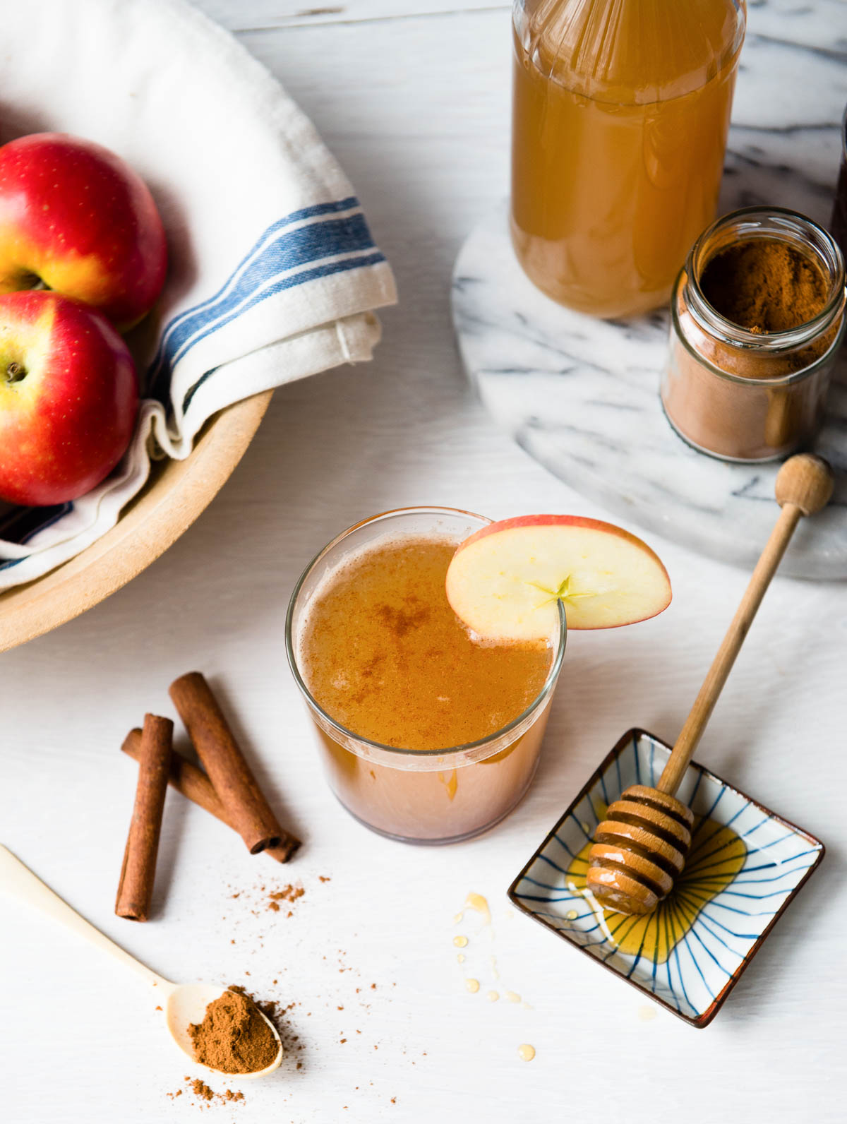 apple cider vinegar drink in a short glass with a apple slice garnish next to a honey comb, cinnamon sticks and a teaspoon of cinnamon.