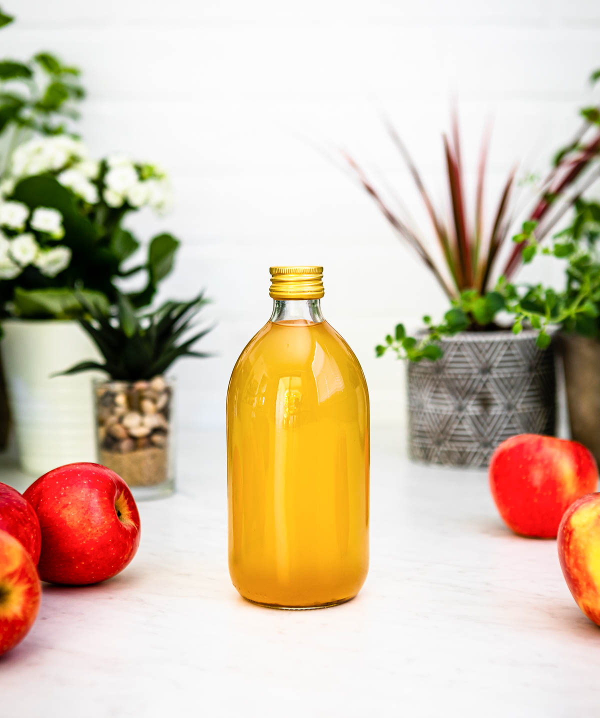 tall glass bottle of apple cider vinegar with a gold lid, sitting on a white counter surrounded by whole apples.
