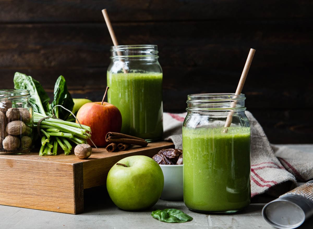 2 glass jars of apple pie smoothie surrounded by fresh apples, cinnamon sticks, nutmeg, spinach and dates.