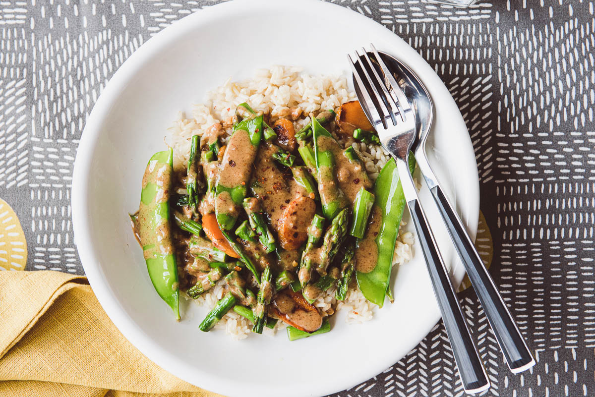 stir-fried asparagus with other veggies on a plate of brown rice and topped with almond butter sauce.