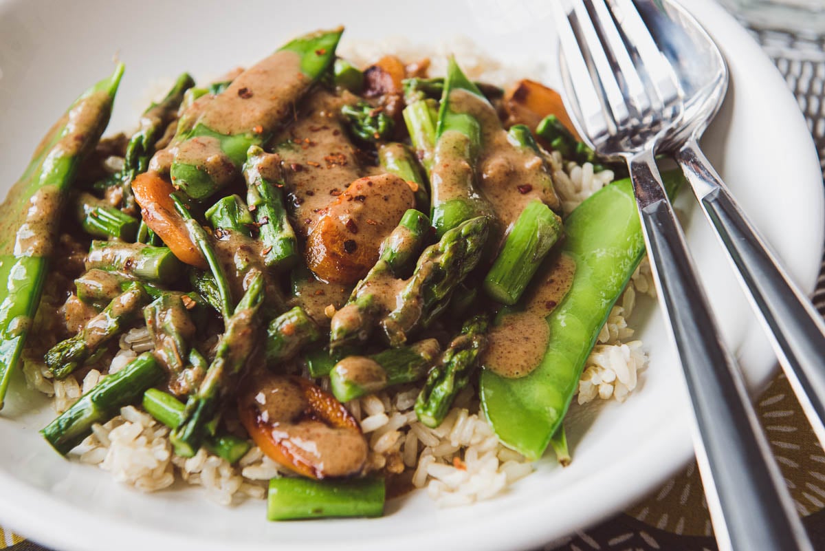white plate of snow peas, asparagus and carrots with brown rice and almond butter sauce.