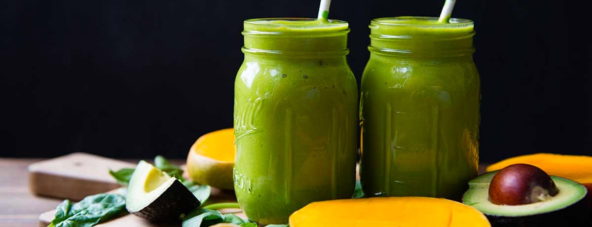 two green smoothies with an halves of avocado sitting next to them