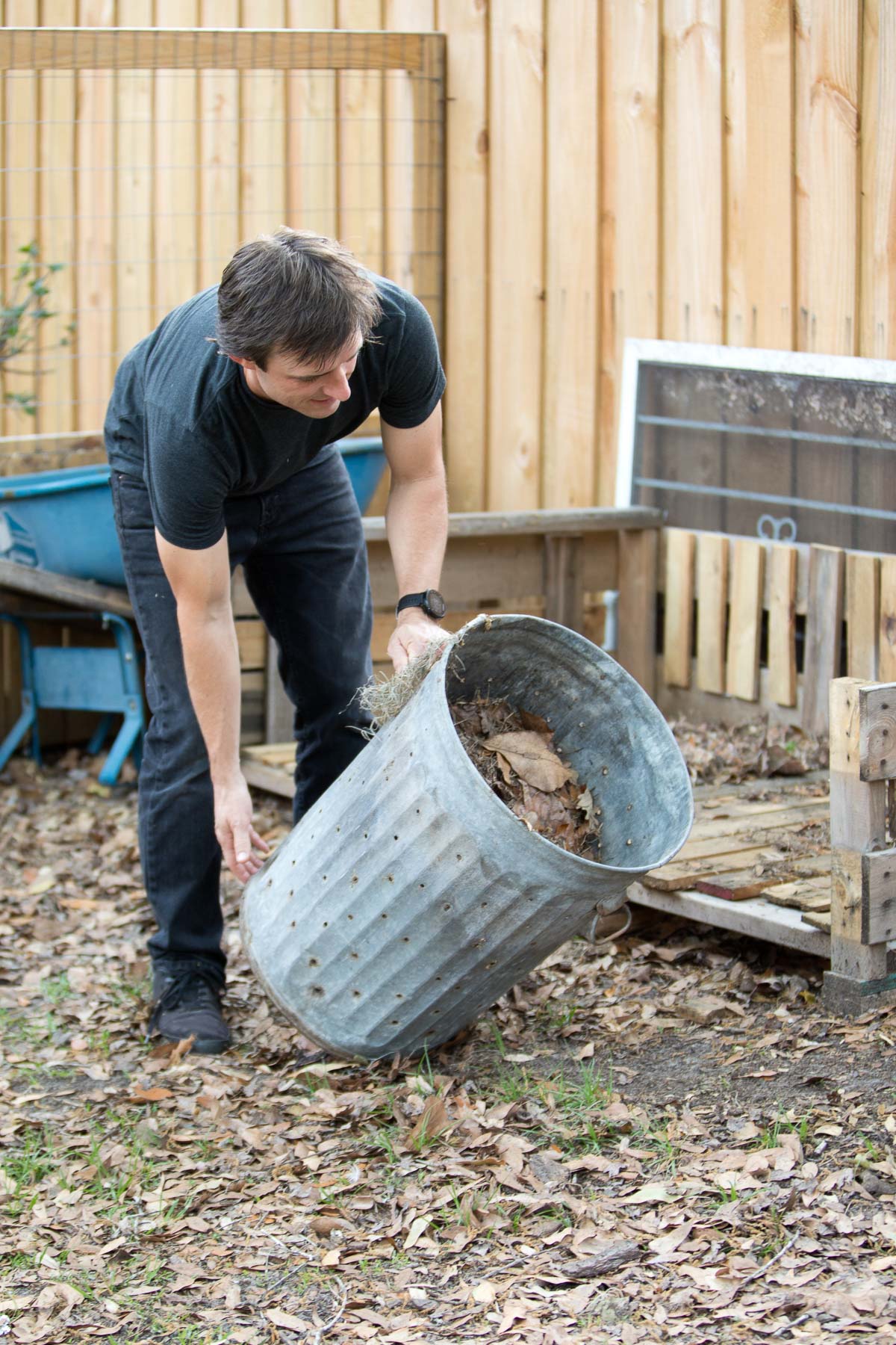 A man lifting a metal trash can filled with compost material to deposit it onto a compost pile.