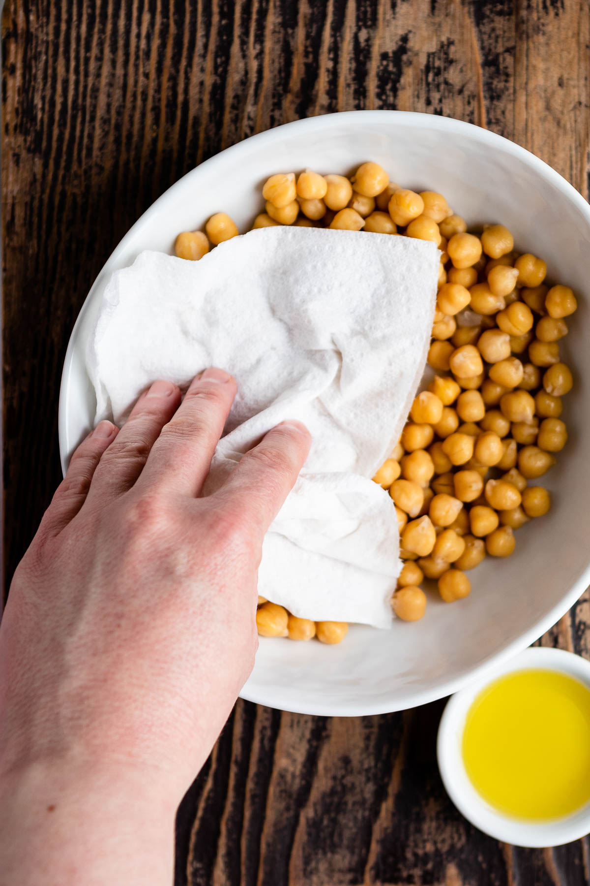 white bowl of garbanzo beans getting dried with a paper towel, next to a small bowl of oil.