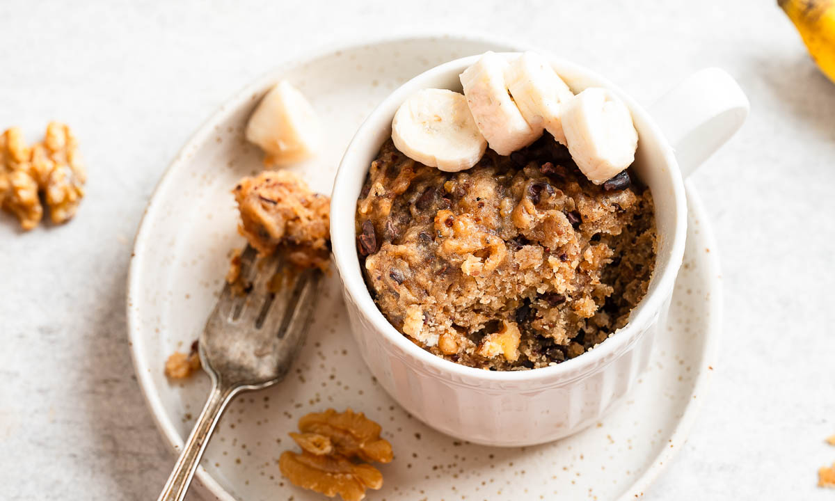 Close-up of a white mug filled with banana cake, topped with sliced banana. 