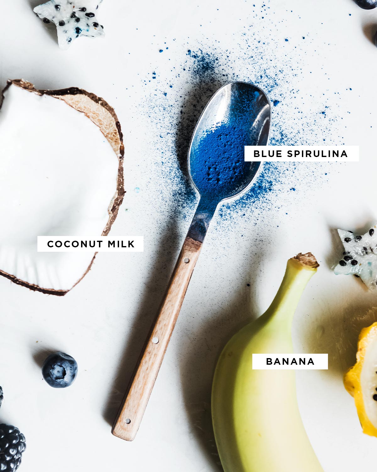 spoon of blue spirulina powder next to a whole banana and cut dragon fruit and coconut.