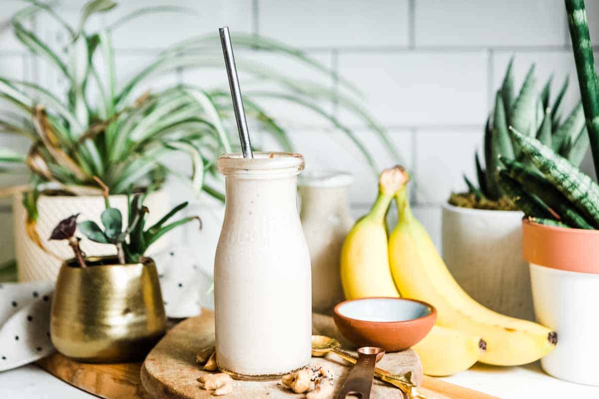 banana smoothie in a glass on the counter with bananas, cashews and measuring spoons.