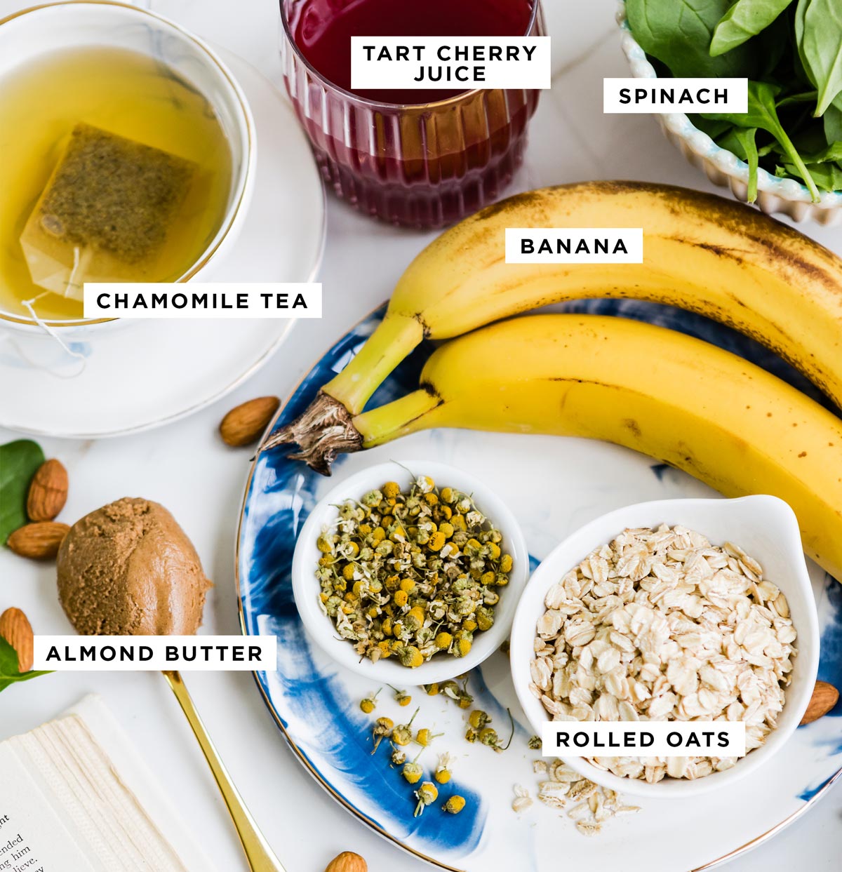 Ingredients for a sleep smoothie recipe, including tart cherry juice, chamomile tea, banana, almond butter, rolled oats, and spinach, arranged on a countertop. 