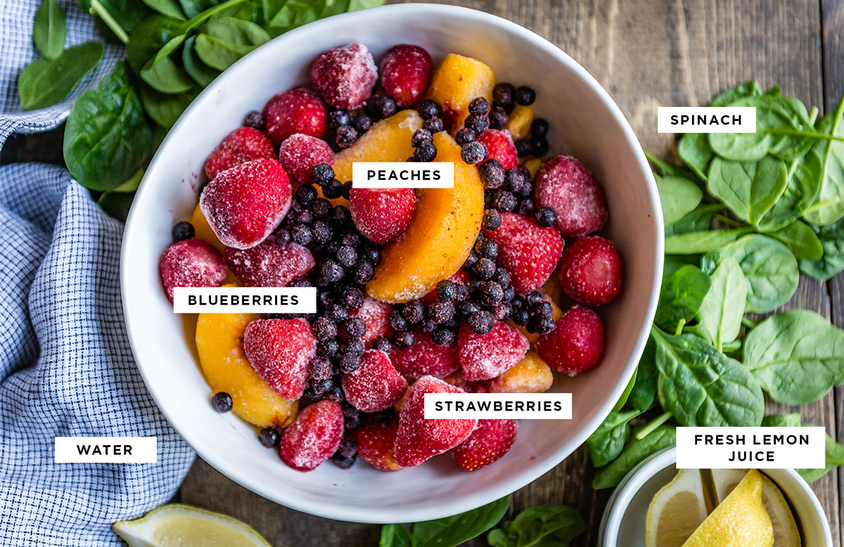 labeled ingredients for a smoothie bowl without banana including spinach, peaches, blueberries, strawberries, water and fresh lemon juice.