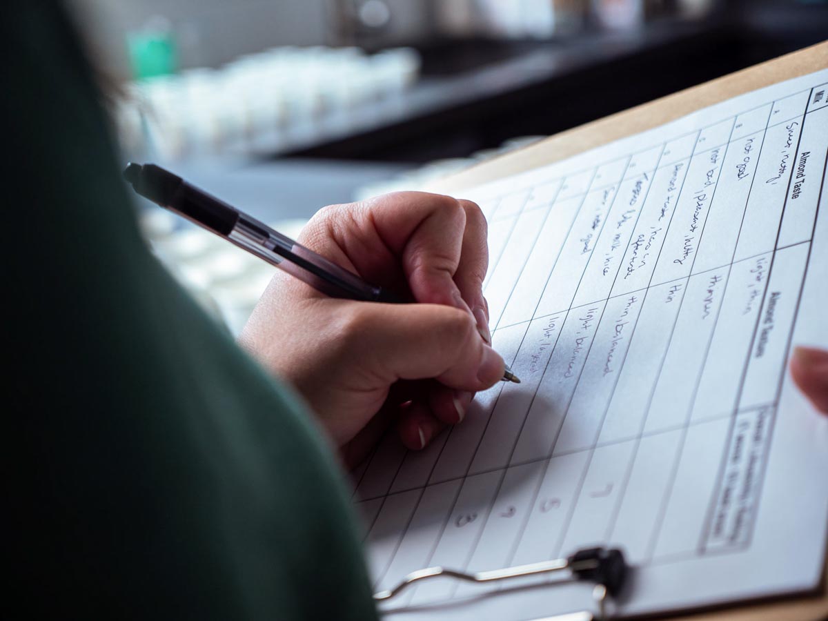 Pen in hand writing score for each almond milk brand on a clipboard.