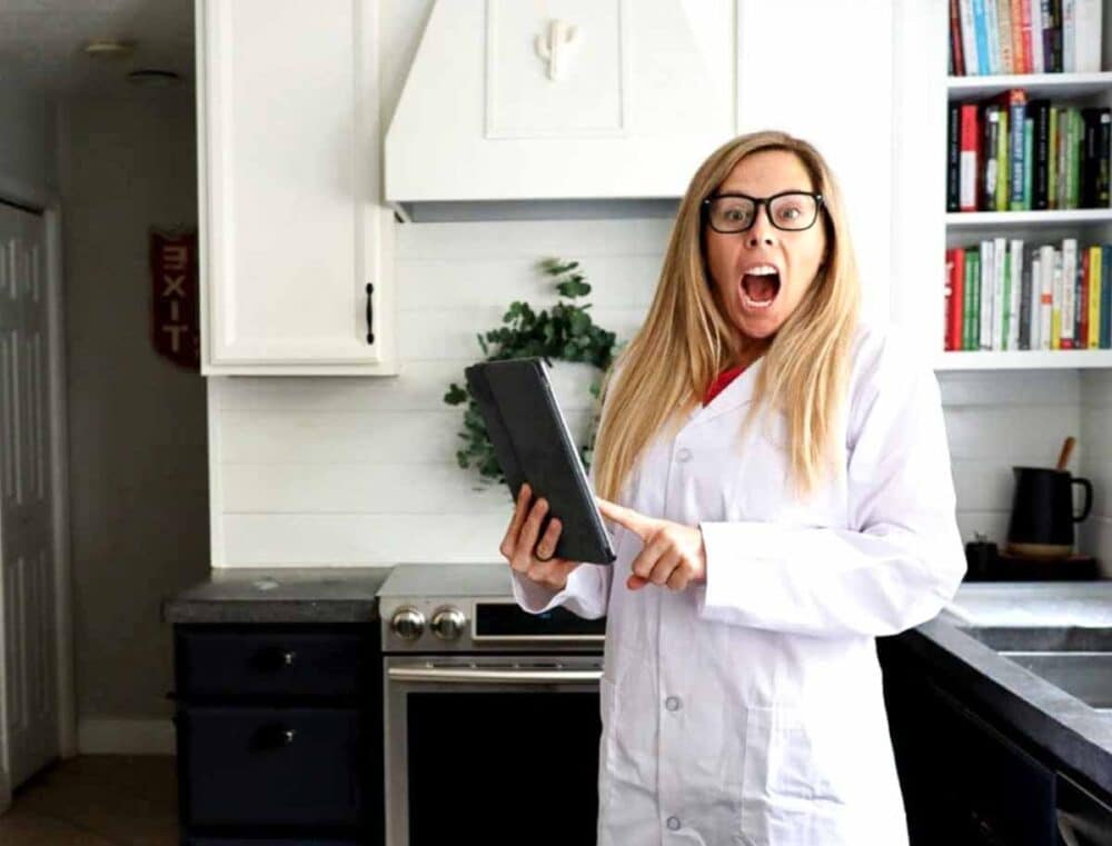 White female with glasses and a lab coat standing in the kitchen holding an iPad, ready to test almonds milk brands.