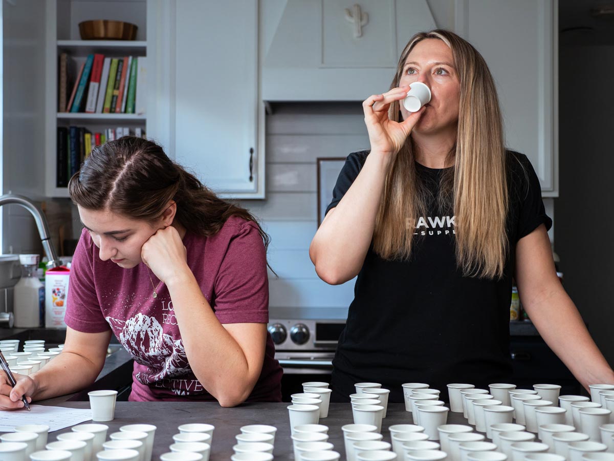 2 white females standing in a kitchen in front of a counter full of dozens of white cups, doing a taste test.