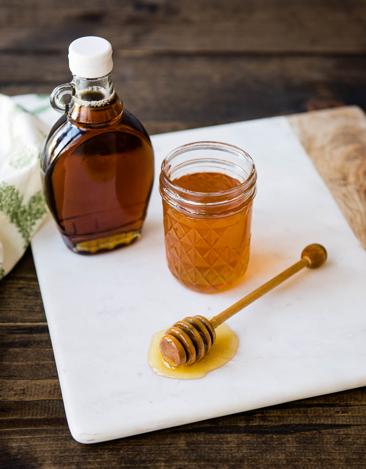 maple syrup and honey both in glass jars and a wooden honey scooper on a white marble cutting board.