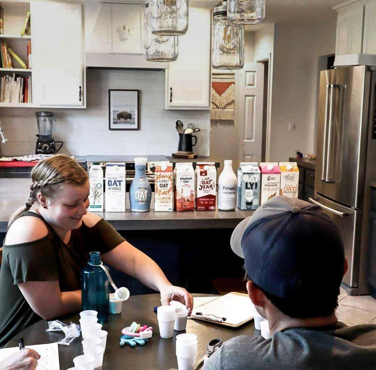 3 people sitting at a table in front of a counter lined with oat milk brands.