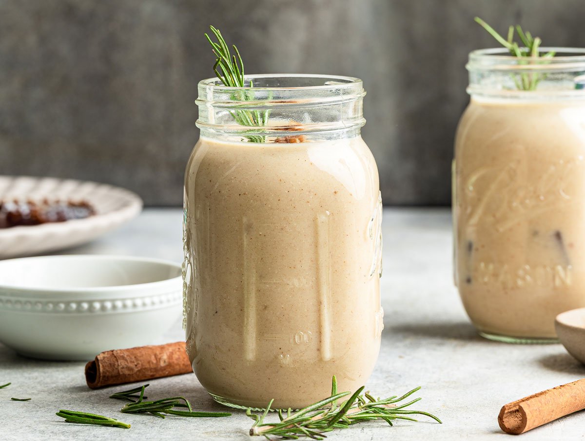 Two vanilla protein shakes on table with cinnamon sticks and rosemary sprig.