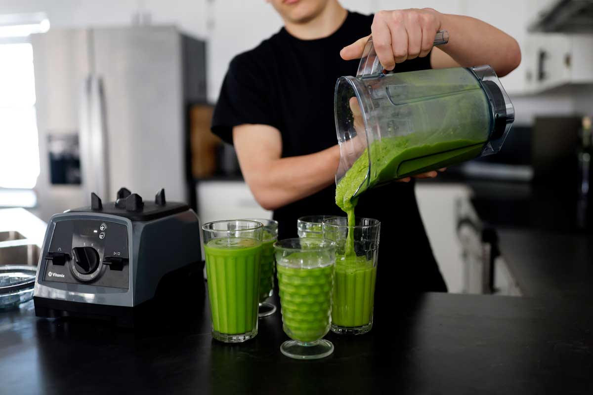 male in black shirt pouring a green smoothies from a Vitamix blender into four glasses in modern kitchen.