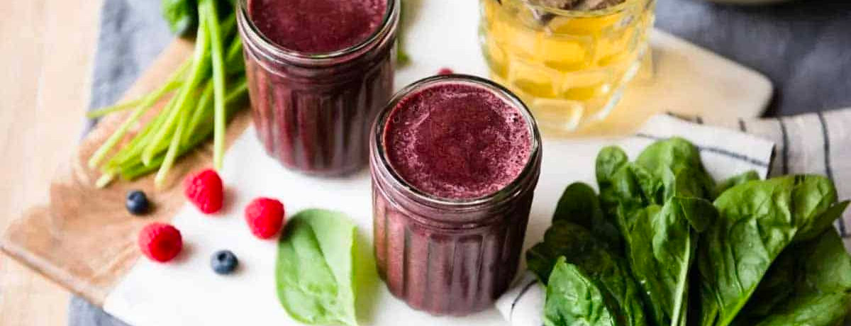 two purple smoothies in glasses with fresh greens and berries on the table in the background