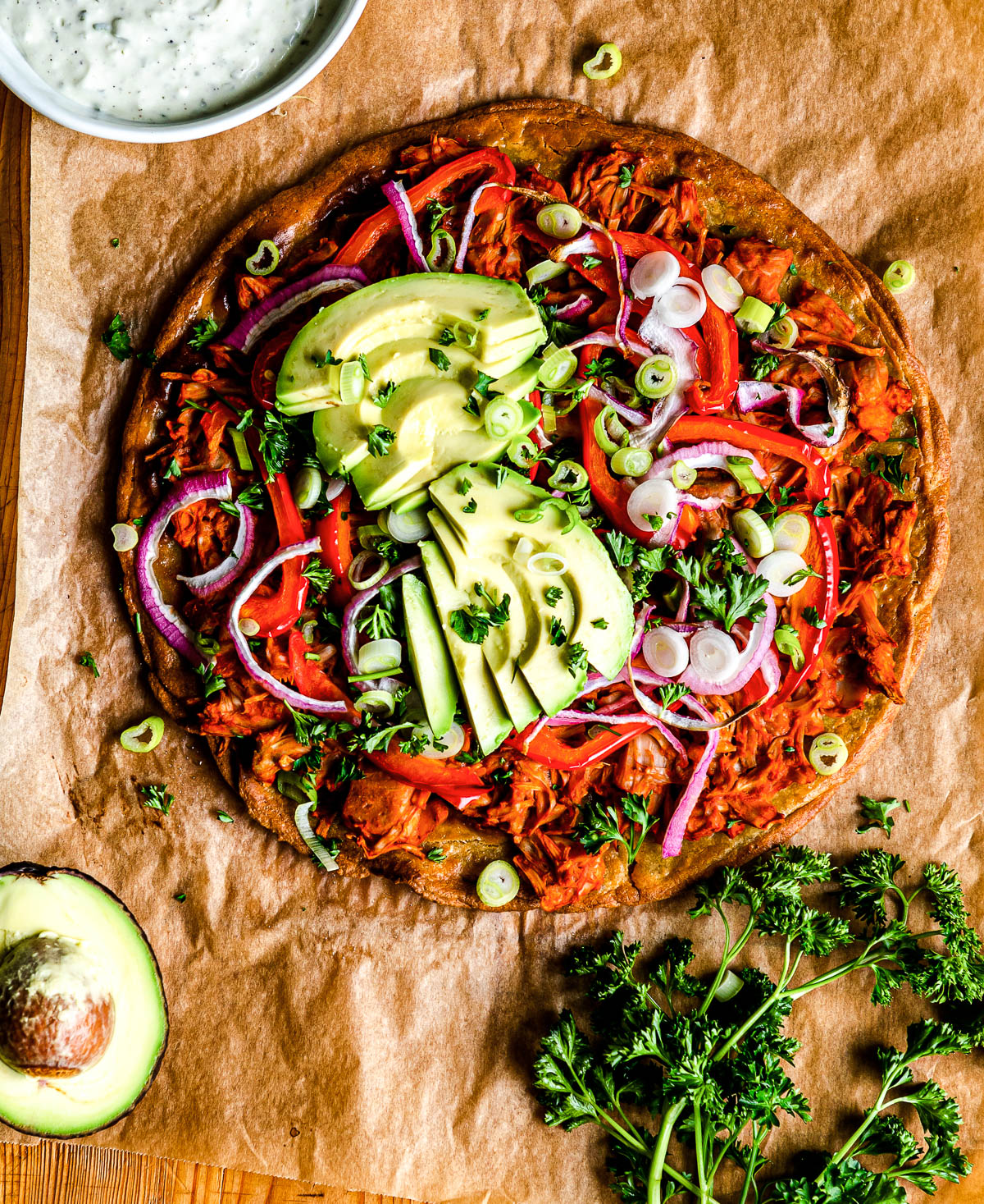 buffalo jackfruit pizza topped with avocado and fresh parsley on parchment paper.