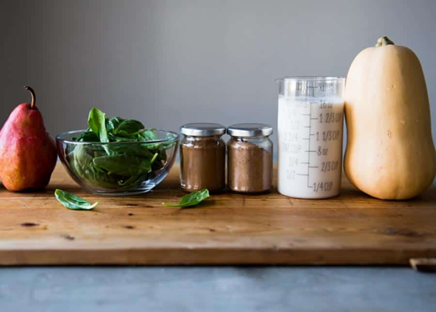 warm smoothie ingredients lined up on a wooden board: pear, spinach, spices, cashew milk and butternut squash.