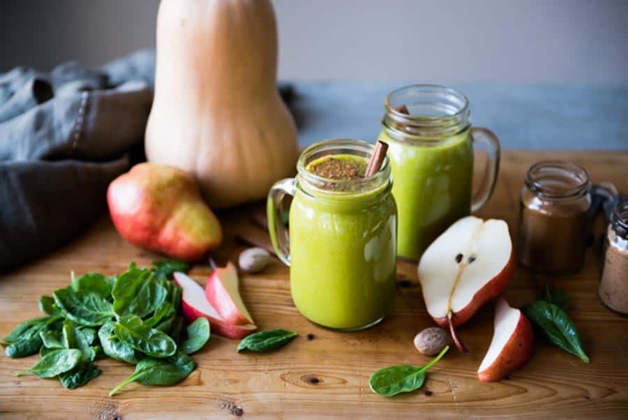 2 glasses of butternut squash smoothie on a wooden board covered with fresh spinach, spices and pears.