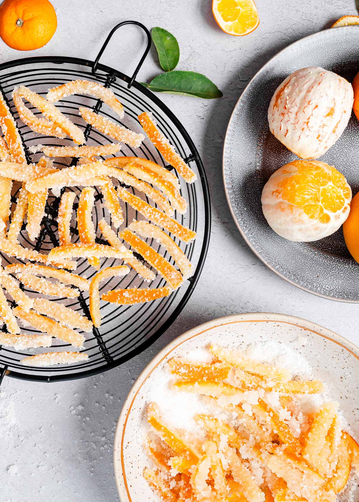 Sugary candied orange peels arranged on a cooling rack next to peeled fresh oranges
