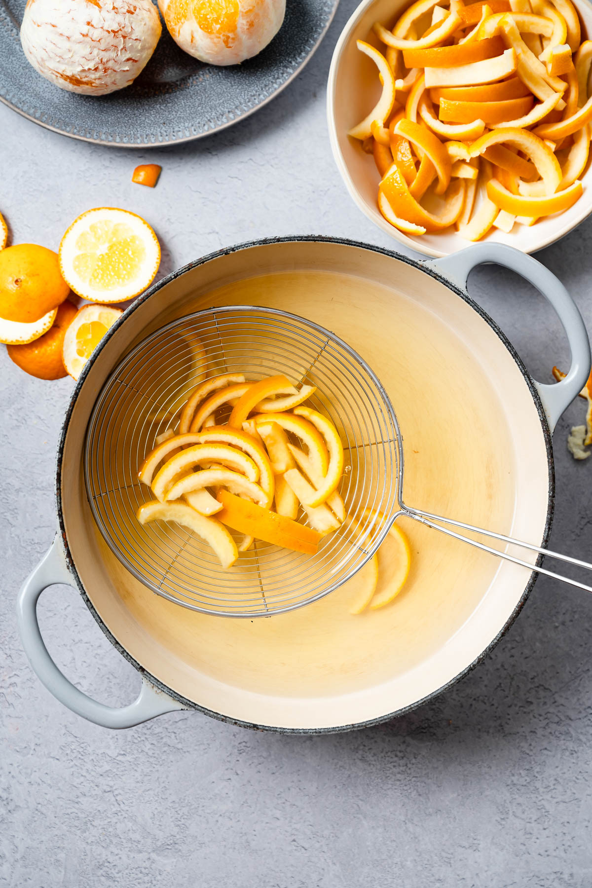 Blanched orange peels being lifted from a pot of hot water with a metal strainer