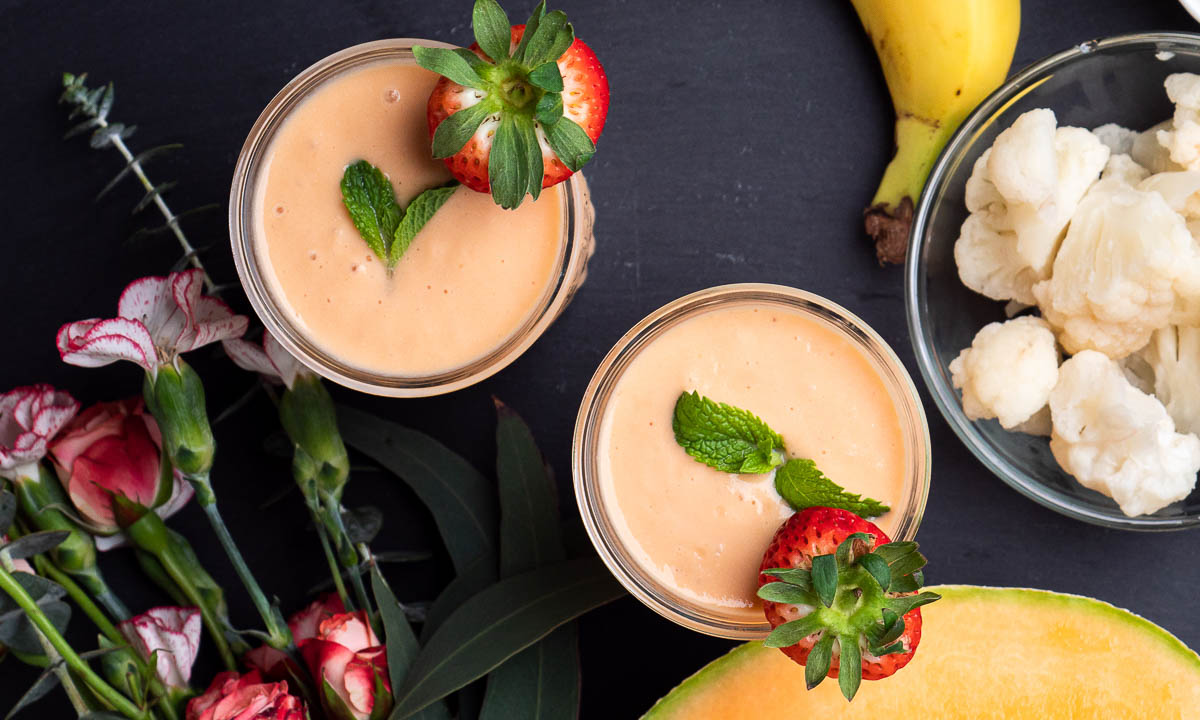 2 orange colored smoothies topped with fresh strawberries and mint leaves, next to flowers, cantaloupe and cauliflower.