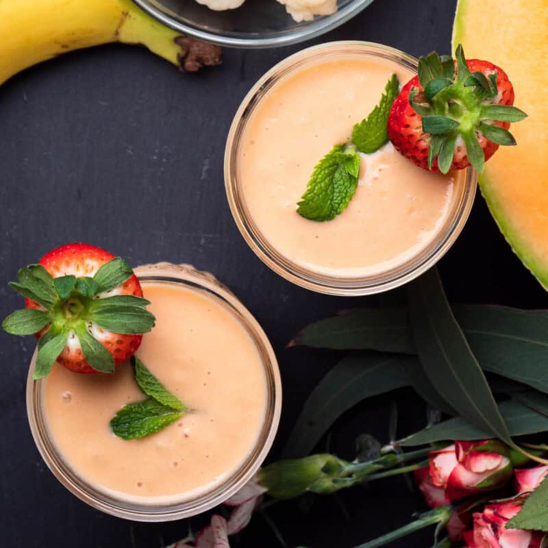 2 glasses with cantaloupe smoothies topped with mint leaves and fresh strawberries.