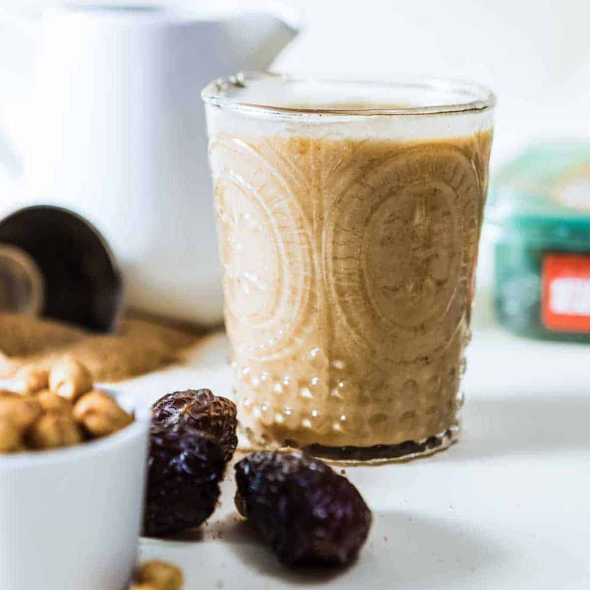 Healthy caramel protein shake in an etched glass next to pitted dates and a white bowl of cashews.