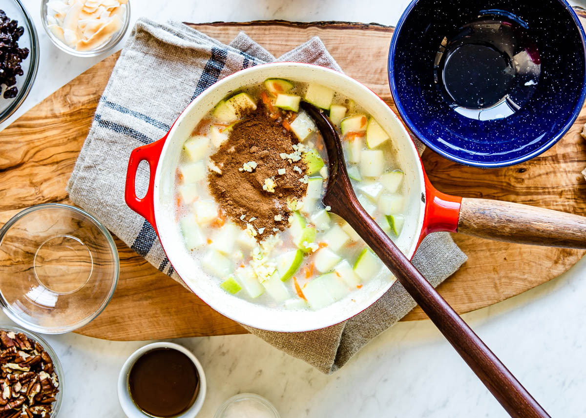 red sauce pan filled with chopped apples, coconut sugar, cinnamon, grated ginger being mixed together with dark wooden spoon.