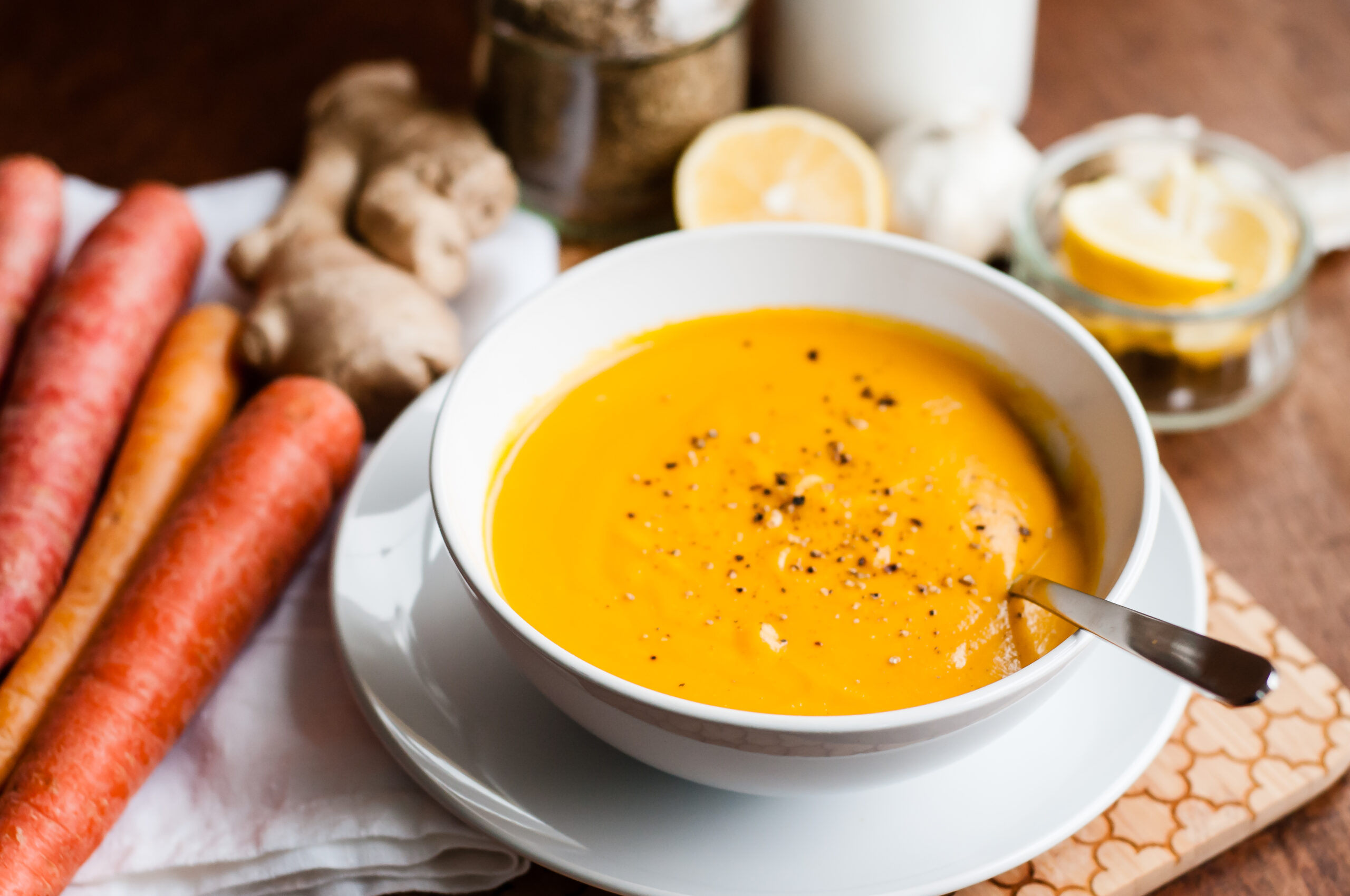 carrot soup in a white bowl on a white plate topped with cracked black pepper and a silver spoon.