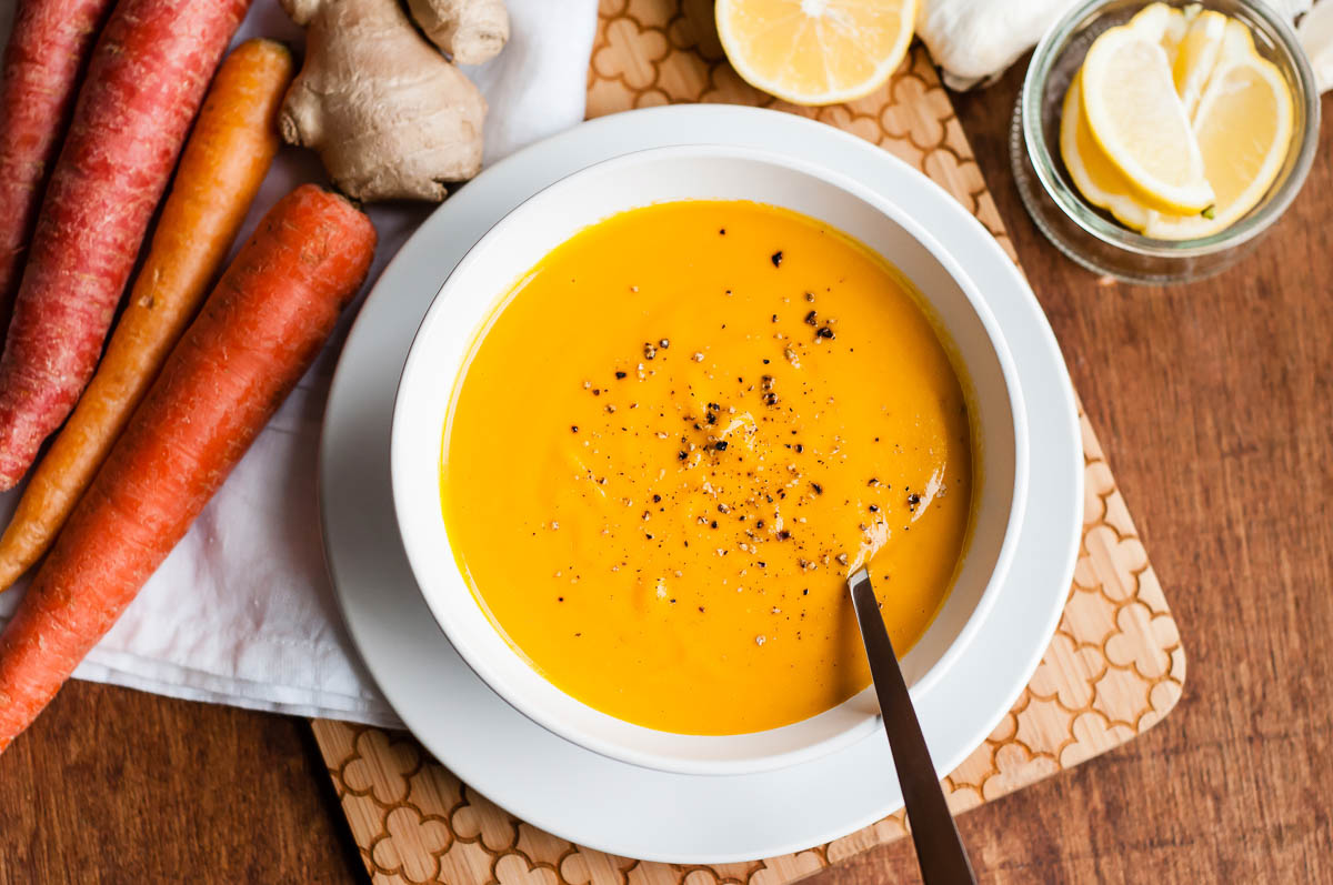 creamy, pureed soup topped with cracked black pepper in a white bowl surrounded by whole carrots, ginger root and sliced lemons.