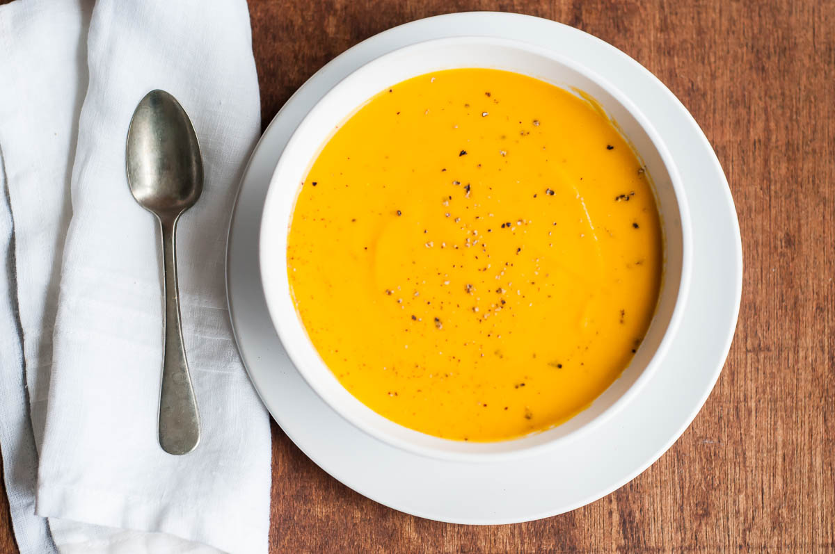 white bowl of carrot ginger soup next to a silver spoon on a white napkin.