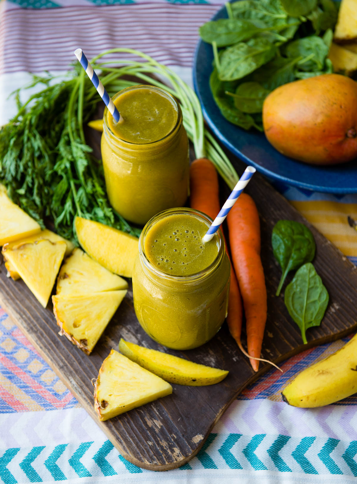 2 glass jars of carrot greens smoothie with papers straws, surrounded by fresh carrots, mango and pineapple slices.