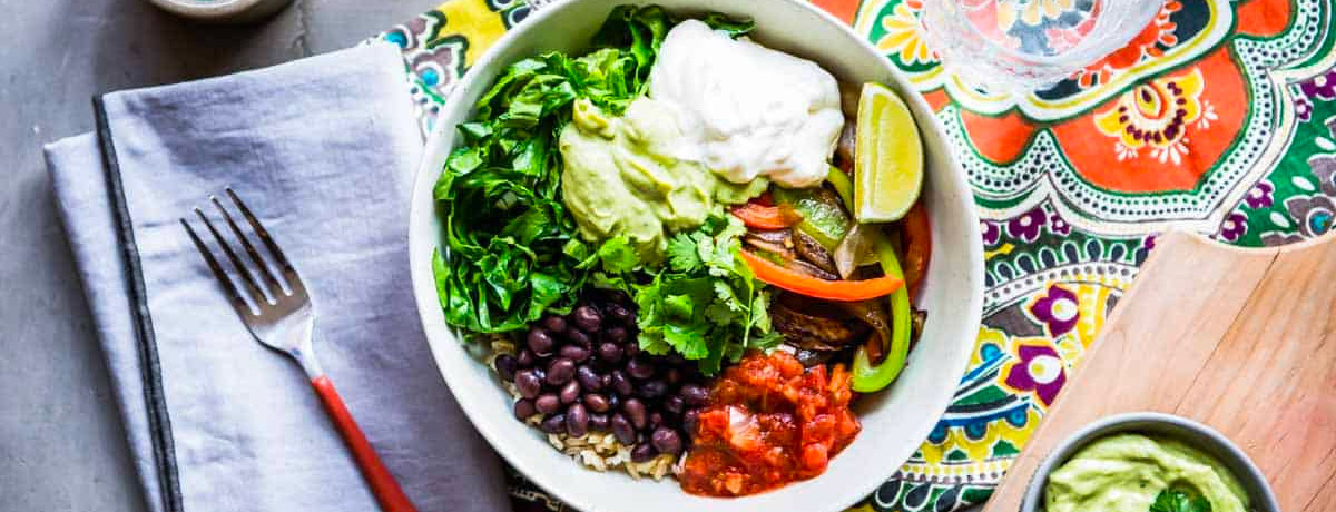 veggie bowl on a table with a napkin, fork, and colorful tablecloth. The bowl has lettuce, black beans, tomatoes, guacamole, crema, and a lime wedge