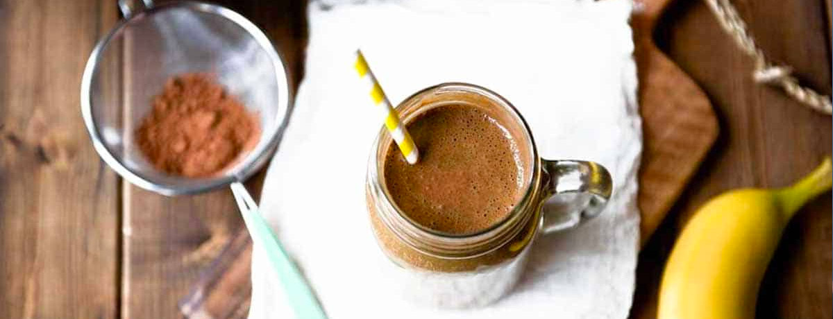 chocolate colored smoothie with a yellow and white striped straw and a sifter with cacao powder in the background