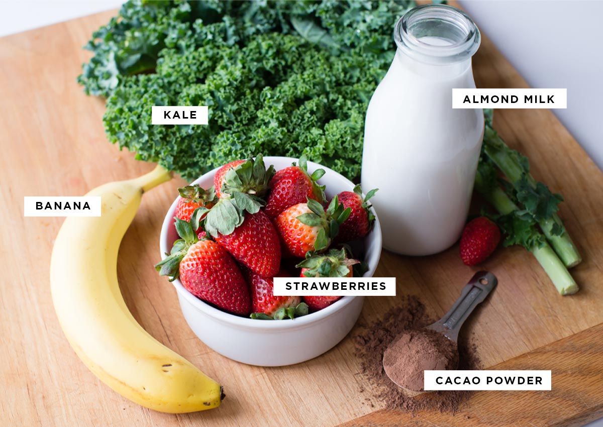 Ingredients for a chocolate strawberry smoothie: kale, banana, strawberries, almond milk, and cacao powder.