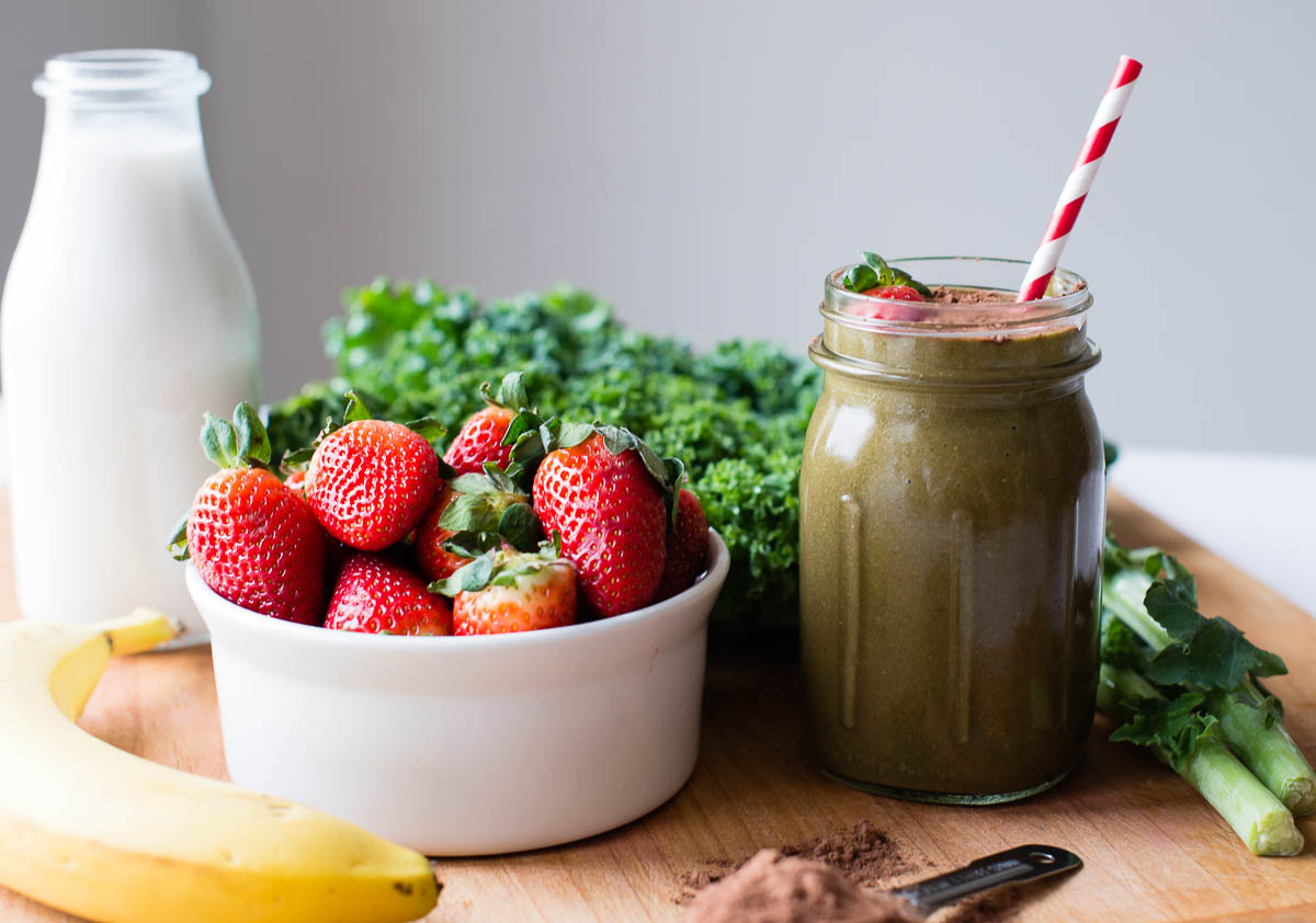 healthy chocolate smoothie in a glass jar next to kale, strawberries, banana and plant-based milk.