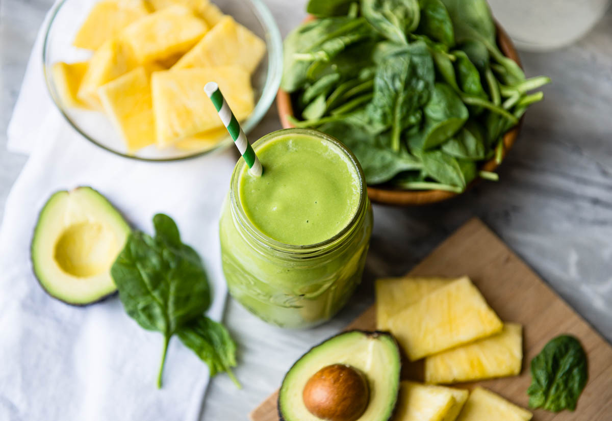 green clear skin smoothie in a mason jar with a paper straw, on a counter with avocado, pineapple and spinach.