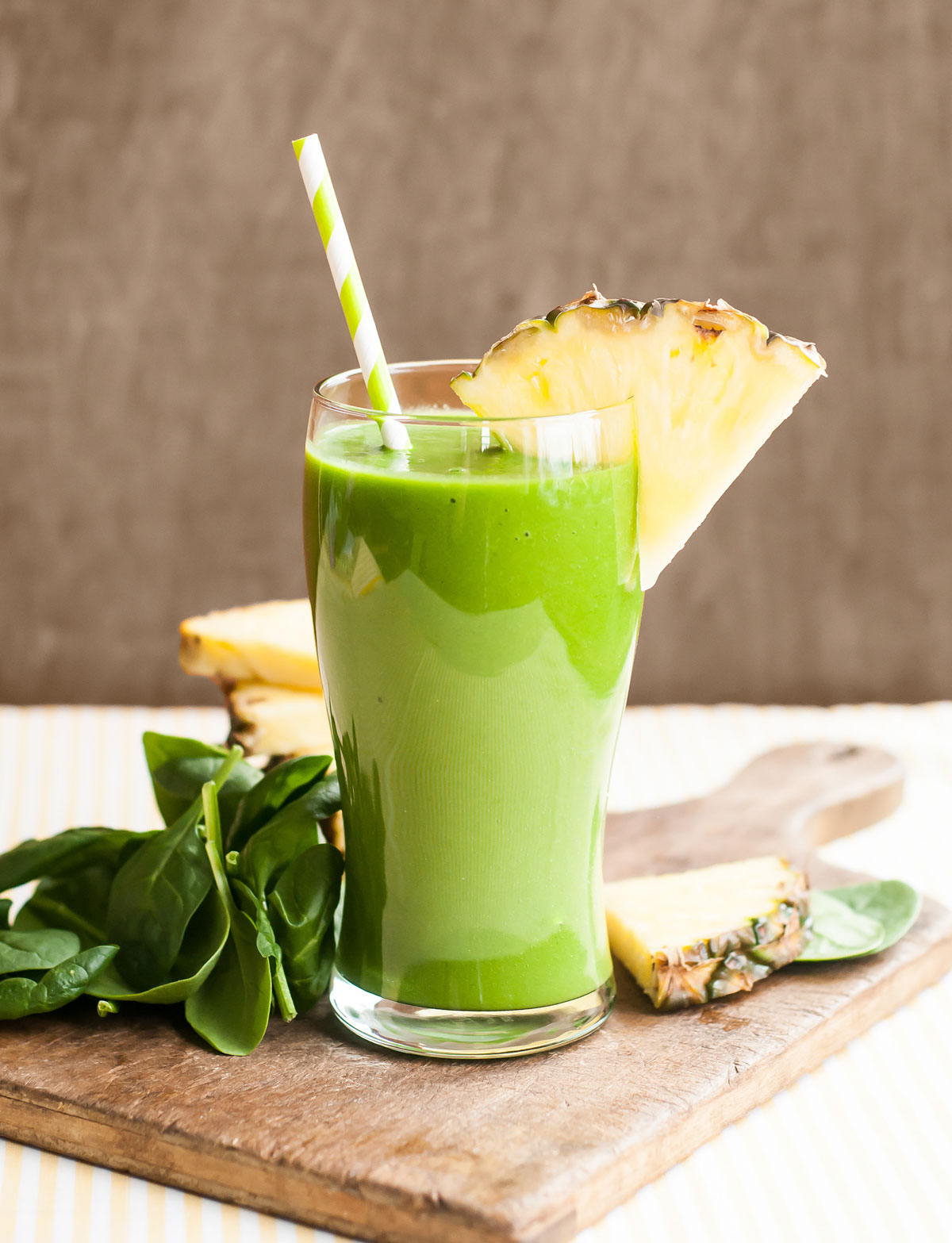 tall glass full of green smoothie with a striped paper straw in it on a wooden cutting board along with spinach and pineapple.