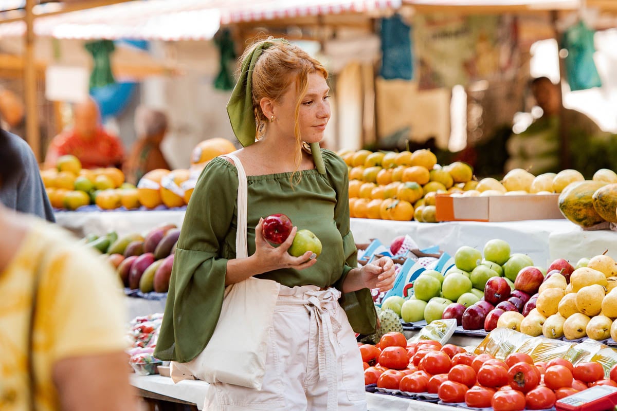 white person with long red hair in a green top shopping for apples at an open market with a canvas tote on their shoulder.