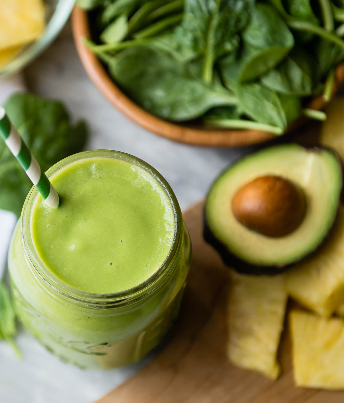 green clear skin smoothie in a mason jar with a paper straw, on a counter with avocado, pineapple and spinach.