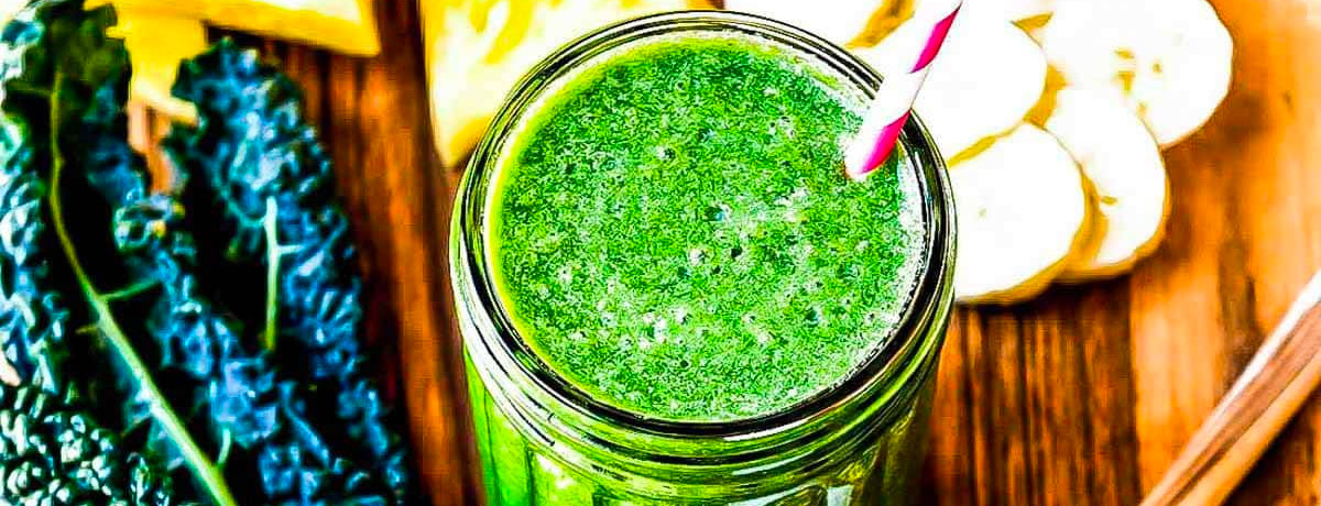 bright green smoothie on a wooden table with ingredients behind it: kale, banana, and the rest are out of focus