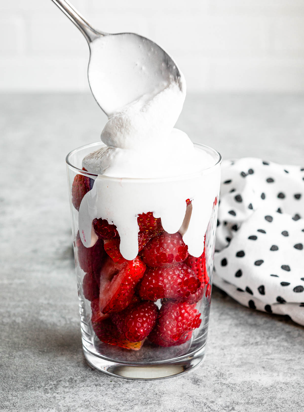 coconut whipped cream getting scooped over top of fresh strawberries in a clear glass.