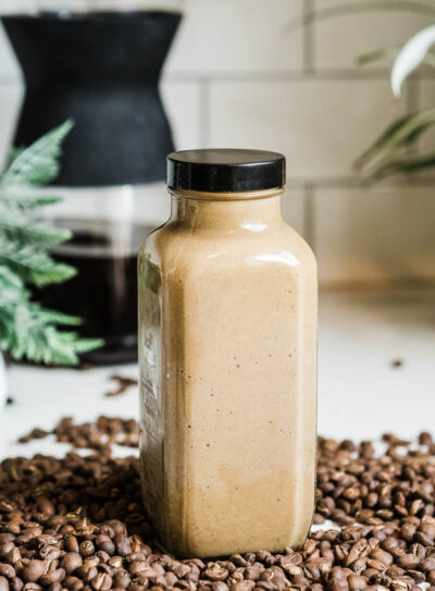 glass container full of a coffee smoothie with a black lid on a countertop full of whole coffee beans.