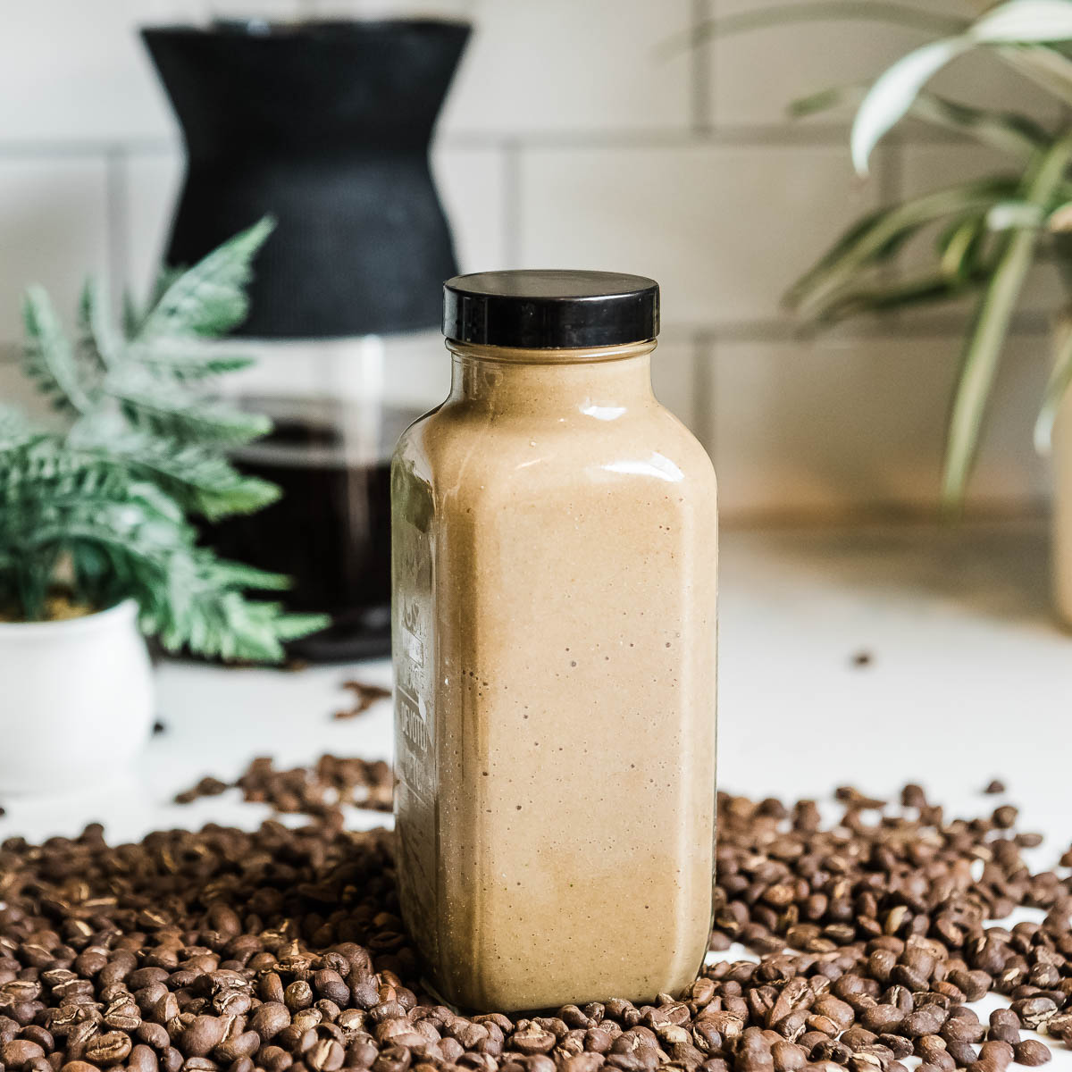 glass container full of a coffee smoothie with a black lid on a countertop full of whole coffee beans.