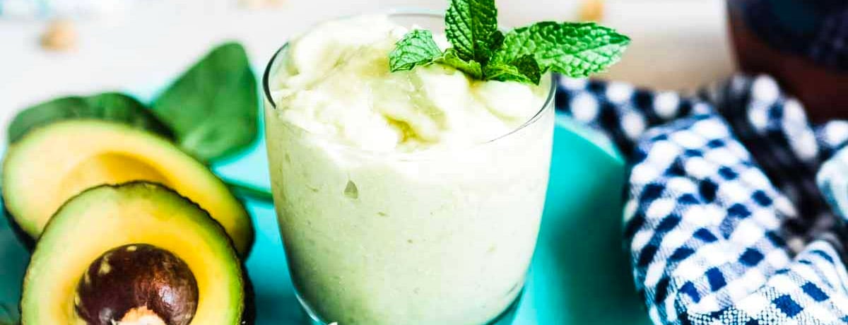 light green colored smoothie with a sprig of mint and halved avocados on a plate next to it