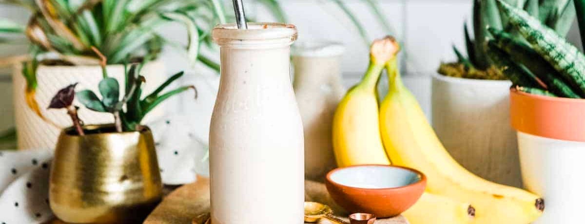 off-white smoothie in a milk jar with bananas and plants in the background
