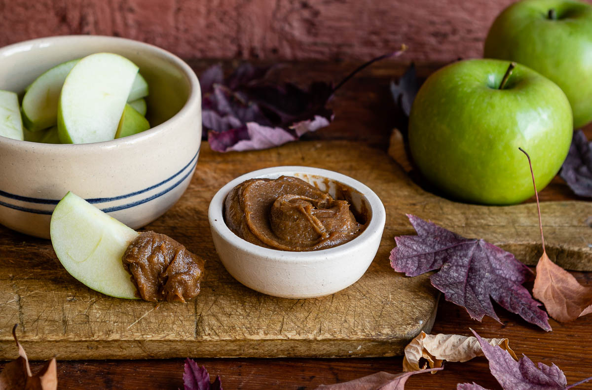 caramel apple dip in a white bowl next to a bowl of sliced green apples on a wooden plank with fake leaves.