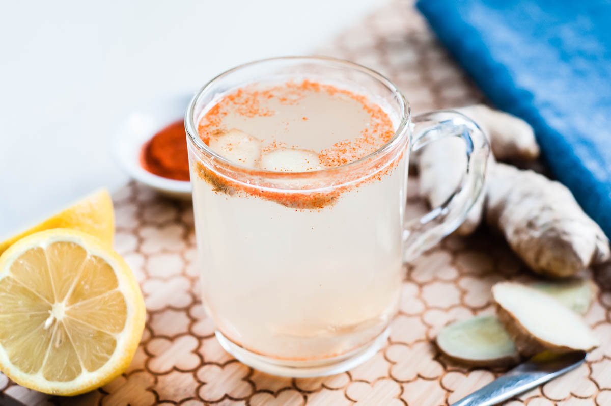 glass mug of cleansing lemon tea topped with cayenne pepper and ginger root on a wooden cutting board surrounded by lemon and ginger.